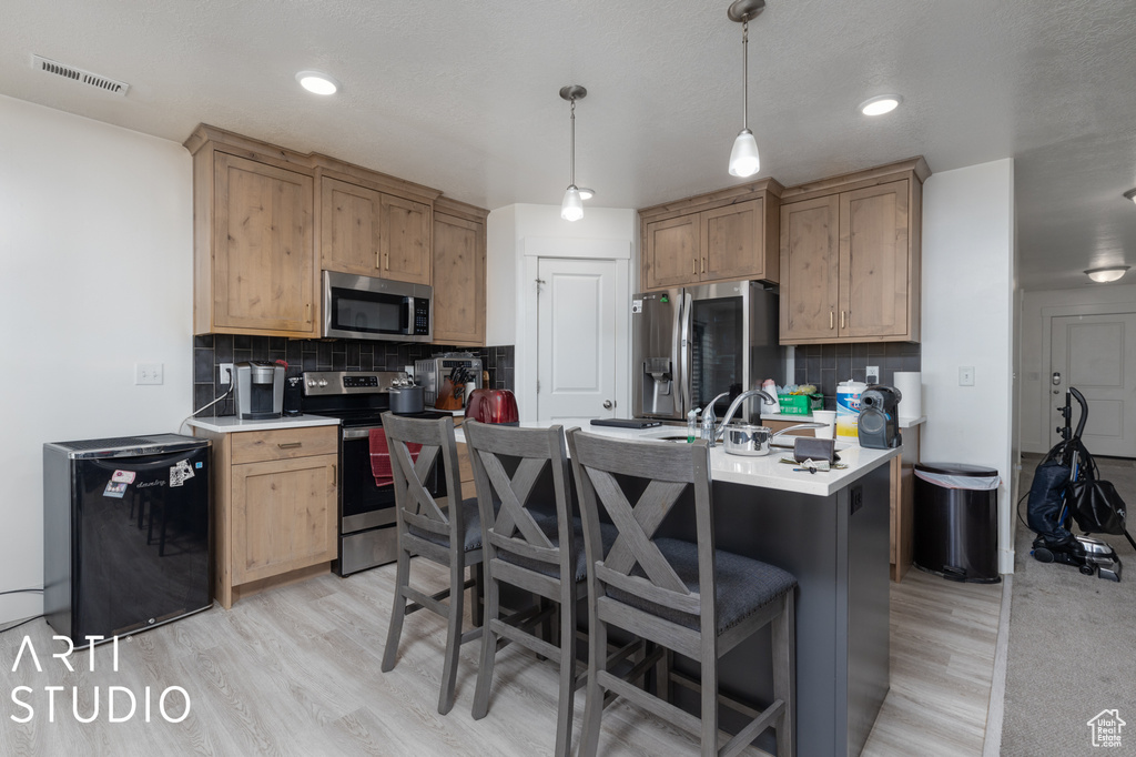 Kitchen featuring backsplash, appliances with stainless steel finishes, a kitchen breakfast bar, hanging light fixtures, and light wood-type flooring