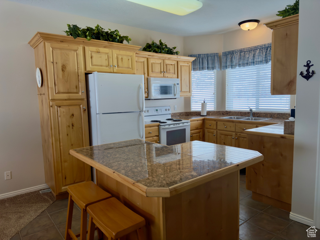 Kitchen featuring light brown cabinetry, dark tile flooring, white appliances, sink, and a kitchen bar