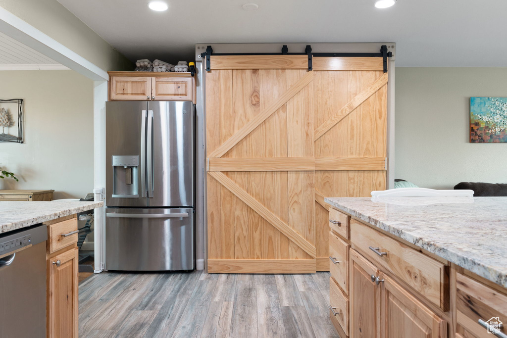 Kitchen featuring light brown cabinetry, light hardwood / wood-style floors, a barn door, appliances with stainless steel finishes, and light stone counters