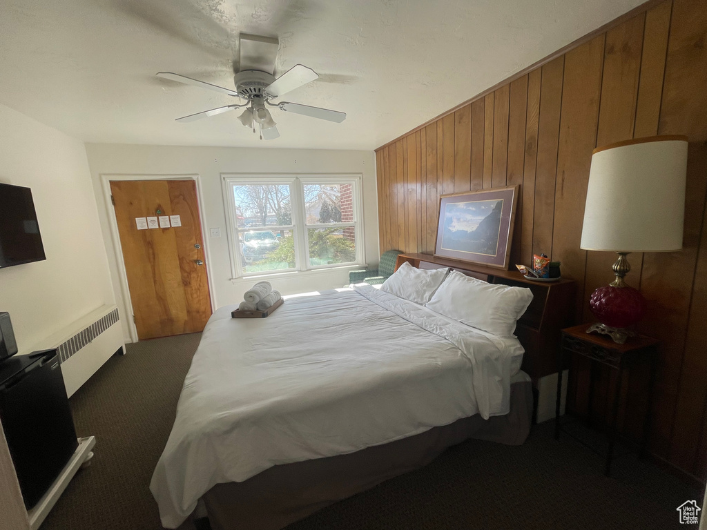 Bedroom with wooden walls, radiator heating unit, dark colored carpet, and ceiling fan