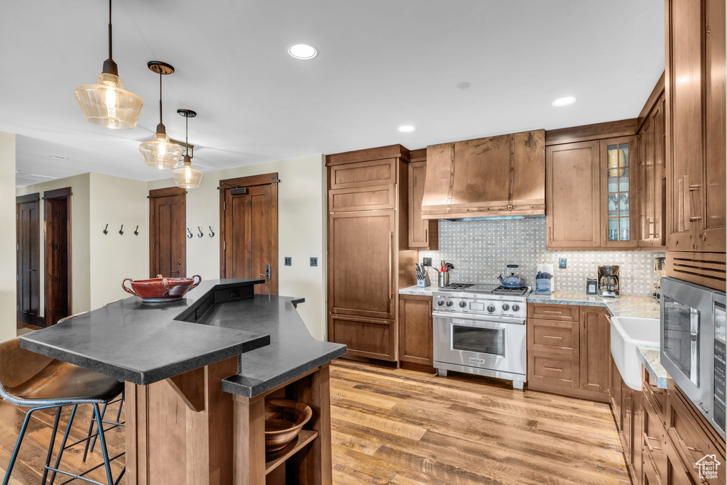Kitchen featuring wall chimney range hood, stainless steel appliances, a kitchen island, light wood-type flooring, and decorative light fixtures