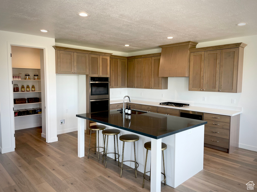 Kitchen featuring double oven, sink, hardwood / wood-style floors, an island with sink, and premium range hood