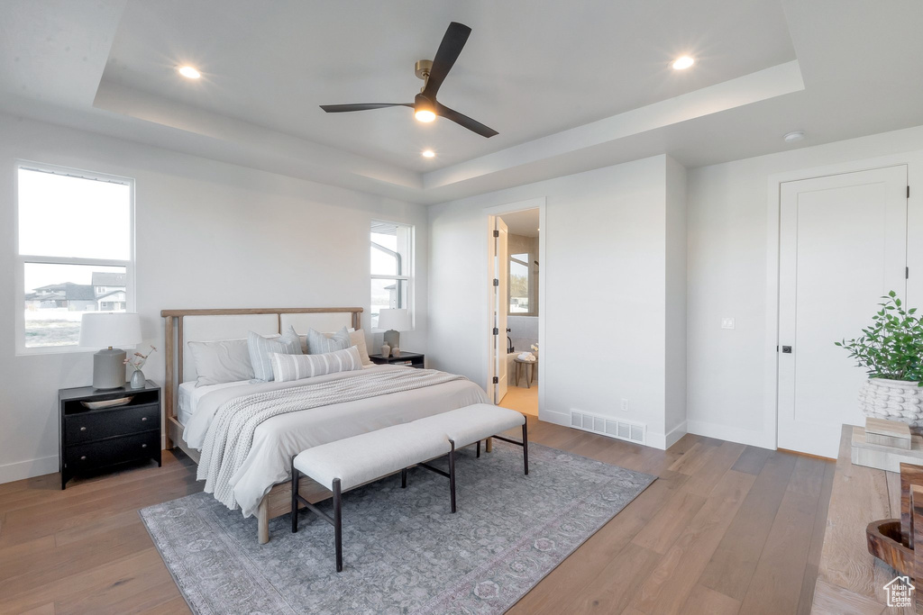 Bedroom featuring ceiling fan, ensuite bathroom, light hardwood / wood-style floors, and a raised ceiling