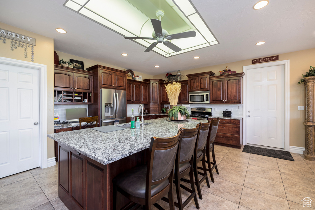 Kitchen with ceiling fan, appliances with stainless steel finishes, a kitchen island with sink, and light stone countertops