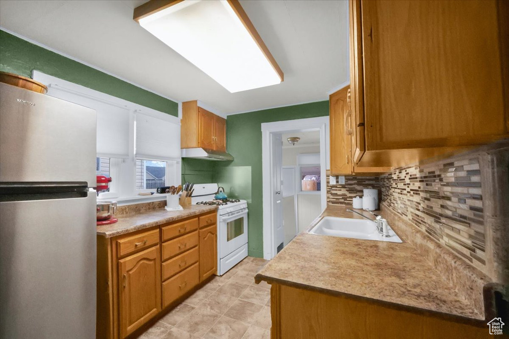 Kitchen with white gas stove, sink, stainless steel refrigerator, backsplash, and light tile floors