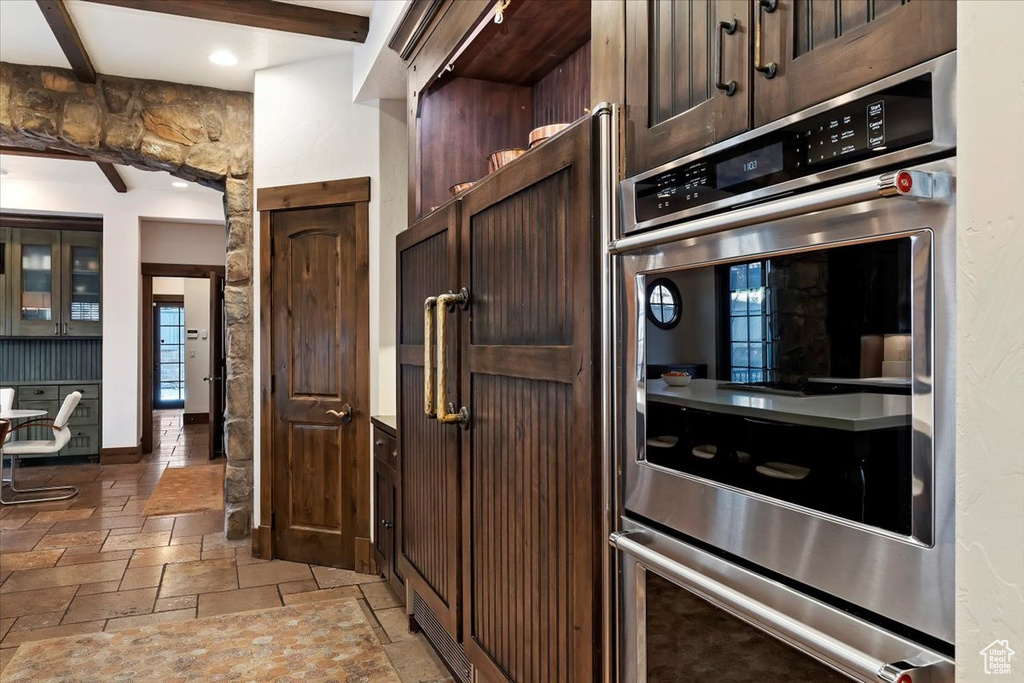 Kitchen featuring light tile flooring, stainless steel double oven, beam ceiling, and dark brown cabinets