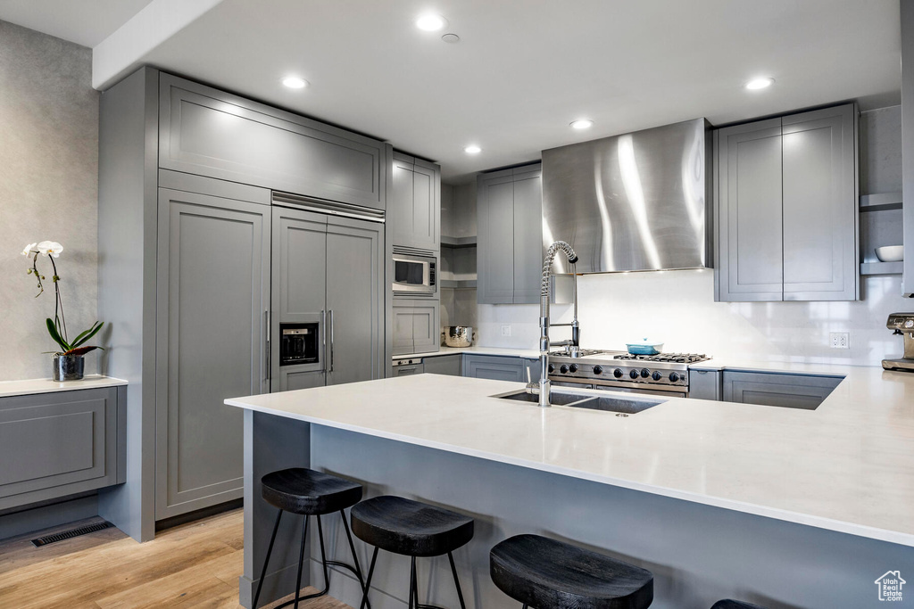 Kitchen featuring wall chimney exhaust hood, a kitchen bar, built in appliances, light hardwood / wood-style floors, and gray cabinets
