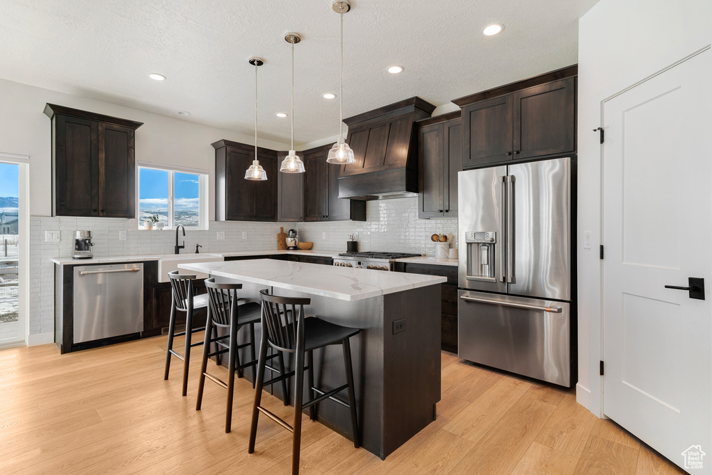 Kitchen with custom exhaust hood, stainless steel appliances, light hardwood / wood-style flooring, decorative light fixtures, and a center island
