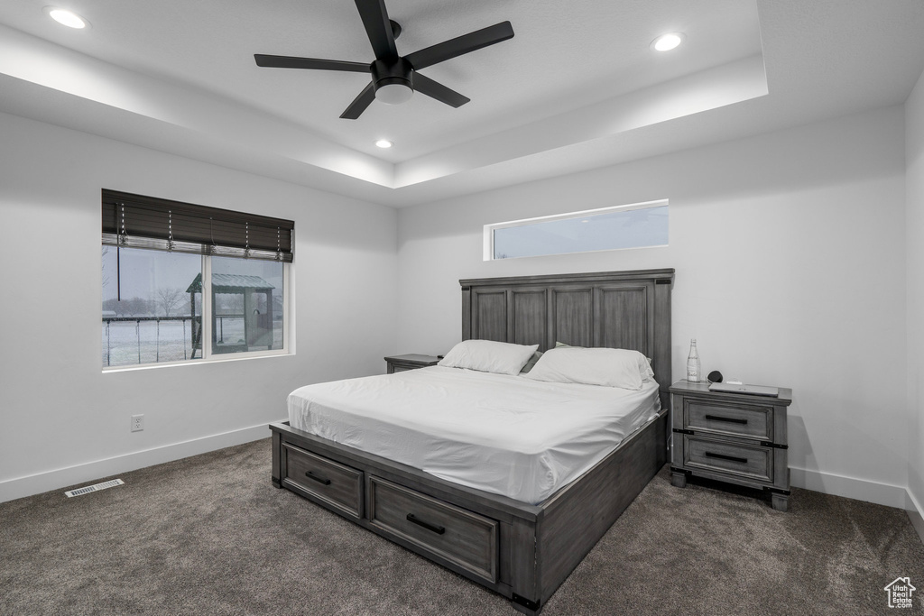 Bedroom with dark carpet, a raised ceiling, multiple windows, and ceiling fan