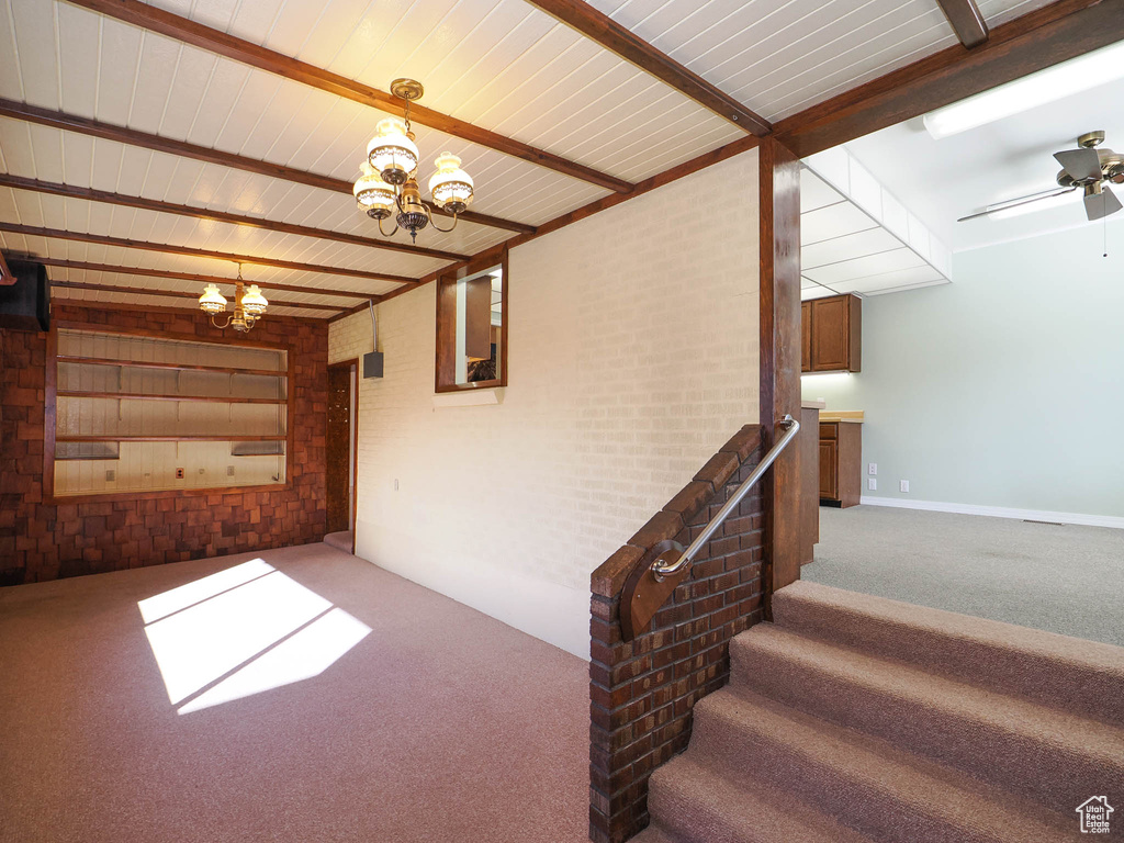 Stairway with dark colored carpet, brick wall, ceiling fan with notable chandelier, and beamed ceiling