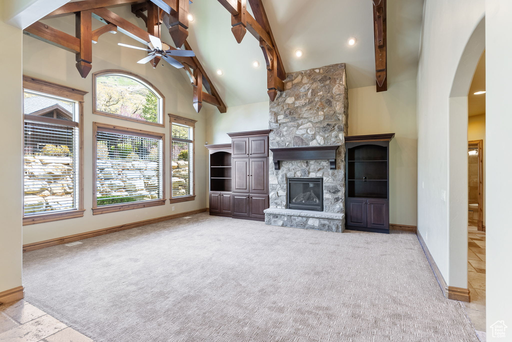 Unfurnished living room featuring ceiling fan, a stone fireplace, high vaulted ceiling, light carpet, and beam ceiling
