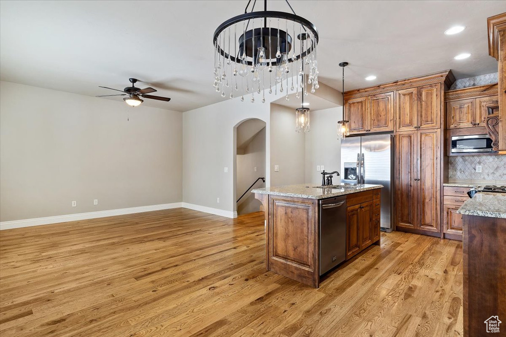 Kitchen featuring a center island with sink, stainless steel appliances, ceiling fan with notable chandelier, hanging light fixtures, and light hardwood / wood-style flooring