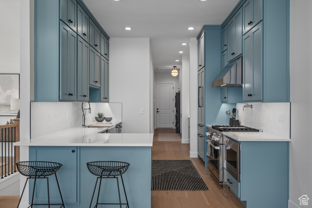 Kitchen featuring backsplash, wall chimney range hood, stainless steel stove, light hardwood / wood-style flooring, and a kitchen bar