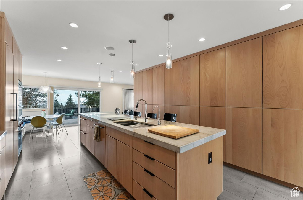 Kitchen with light brown cabinetry, sink, a center island with sink, light tile floors, and pendant lighting