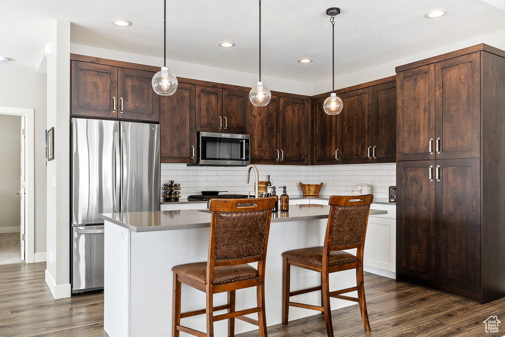 Kitchen featuring dark hardwood / wood-style floors, pendant lighting, tasteful backsplash, a center island with sink, and stainless steel appliances