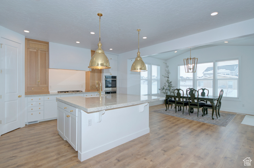 Kitchen with white cabinets, lofted ceiling, hanging light fixtures, an island with sink, and light wood-type flooring