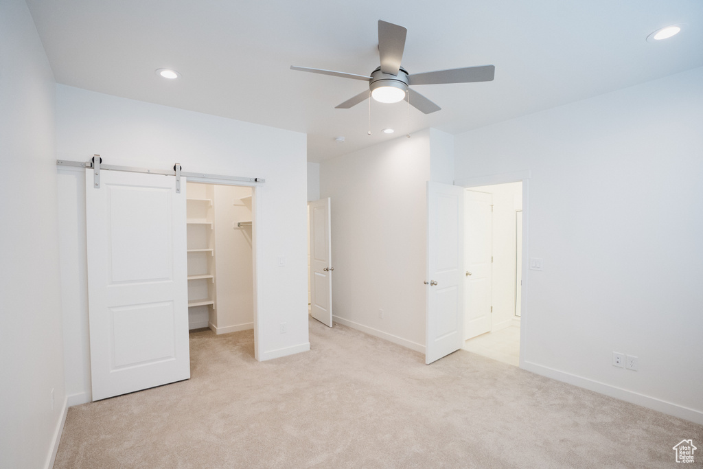 Unfurnished bedroom with light colored carpet, a spacious closet, a barn door, ceiling fan, and a closet