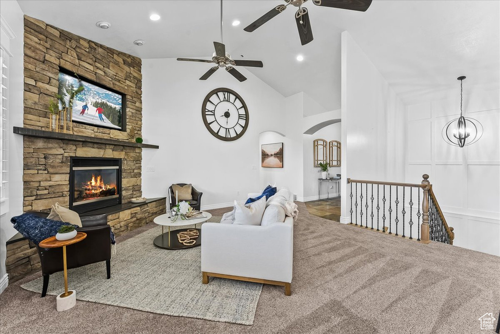 Living room featuring ceiling fan with notable chandelier, a stone fireplace, light colored carpet, and high vaulted ceiling