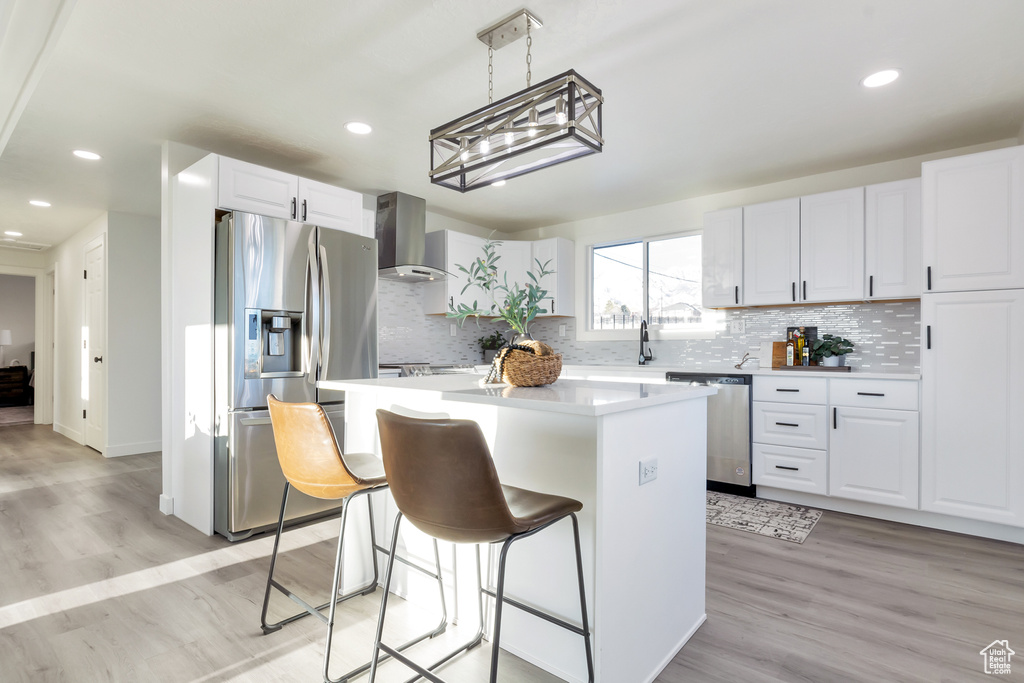 Kitchen featuring a kitchen island, wall chimney exhaust hood, appliances with stainless steel finishes, light hardwood / wood-style flooring, and white cabinetry