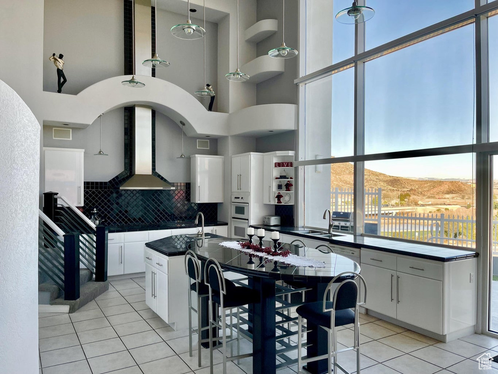 Kitchen featuring white cabinets, a towering ceiling, wall chimney range hood, and decorative light fixtures