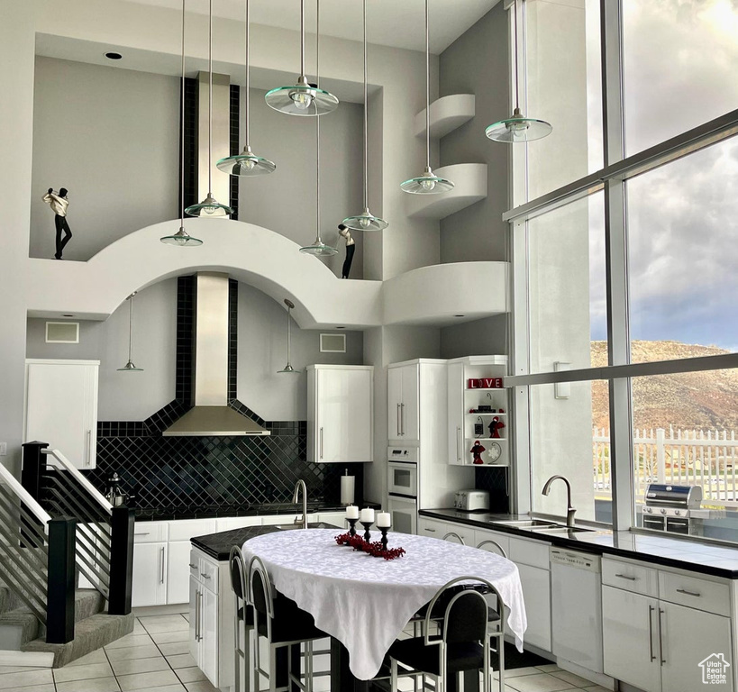 Kitchen with white appliances, wall chimney exhaust hood, a high ceiling, backsplash, and light tile floors