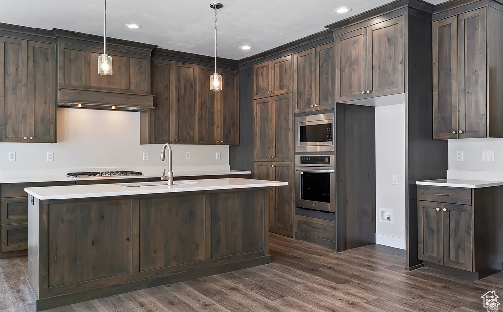 Kitchen with hanging light fixtures, dark hardwood / wood-style flooring, a center island with sink, stainless steel appliances, and dark brown cabinets