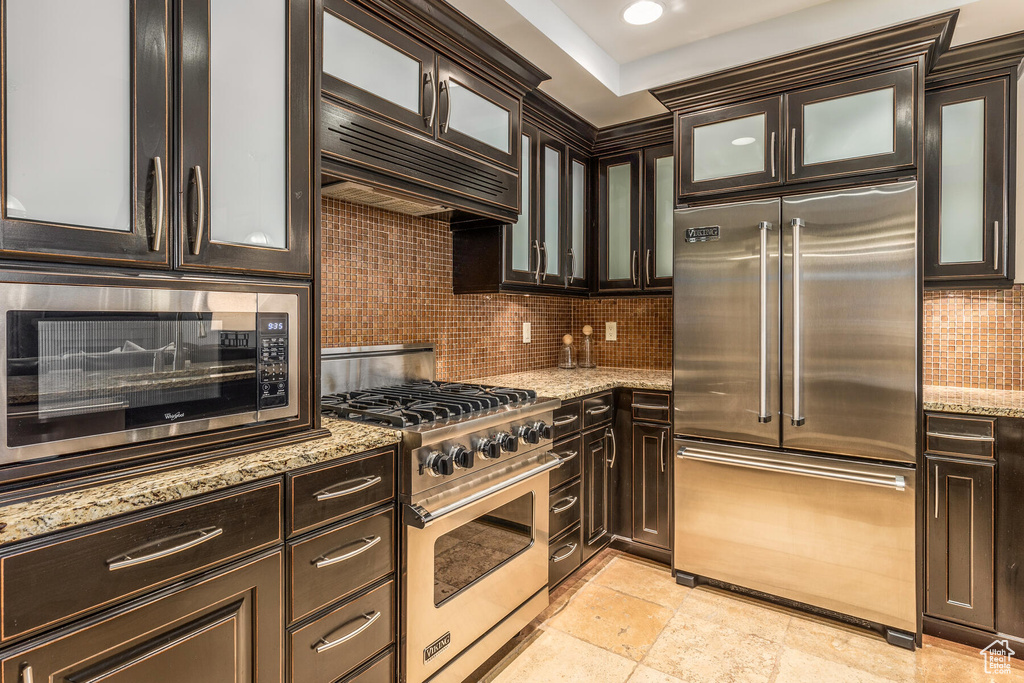 Kitchen with tasteful backsplash, premium appliances, custom exhaust hood, light stone counters, and dark brown cabinetry