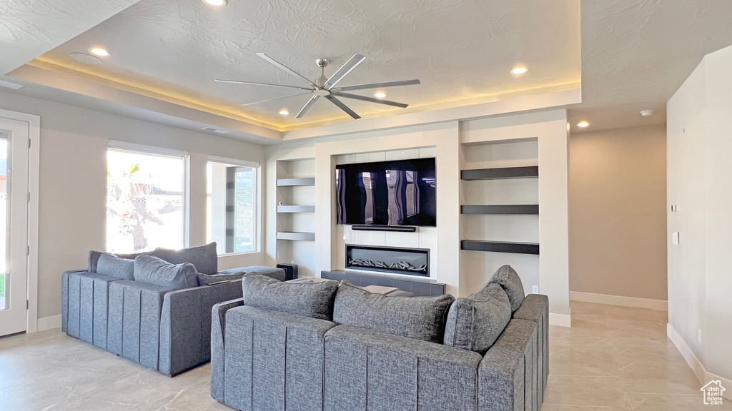Living room with light tile flooring, a raised ceiling, a textured ceiling, built in shelves, and ceiling fan