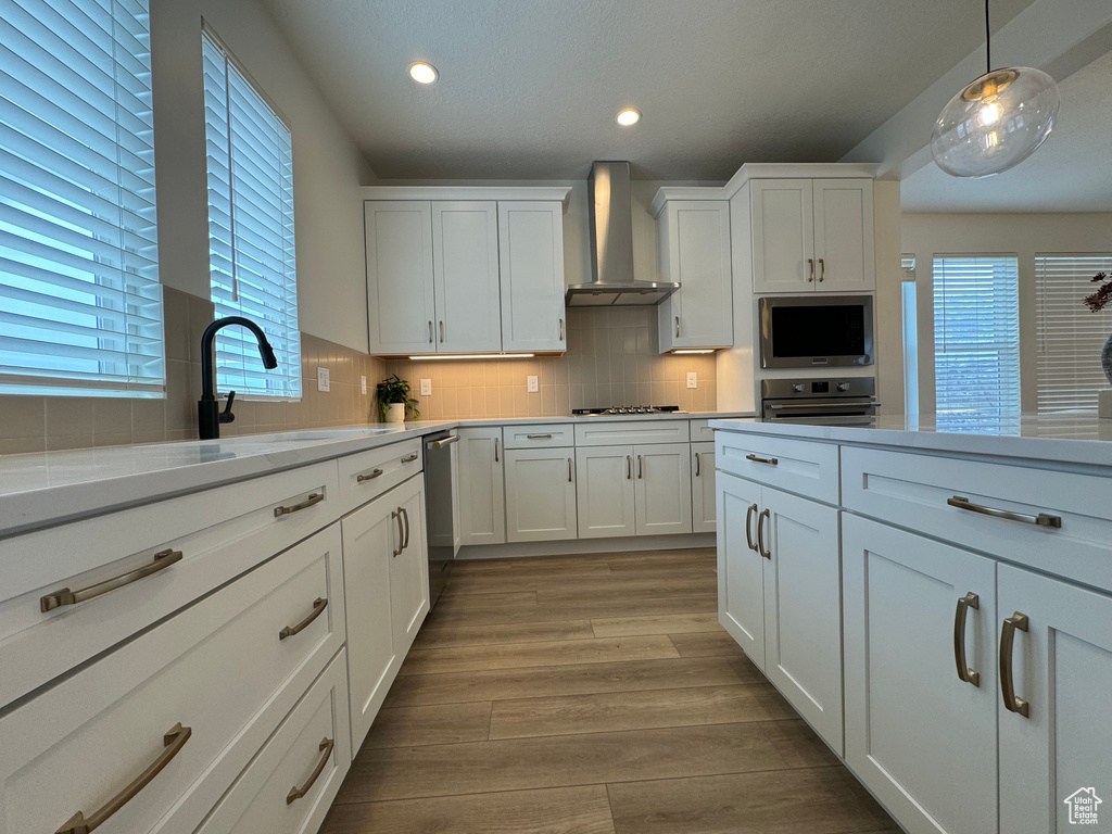Kitchen featuring wall chimney range hood, pendant lighting, tasteful backsplash, appliances with stainless steel finishes, and light hardwood / wood-style flooring