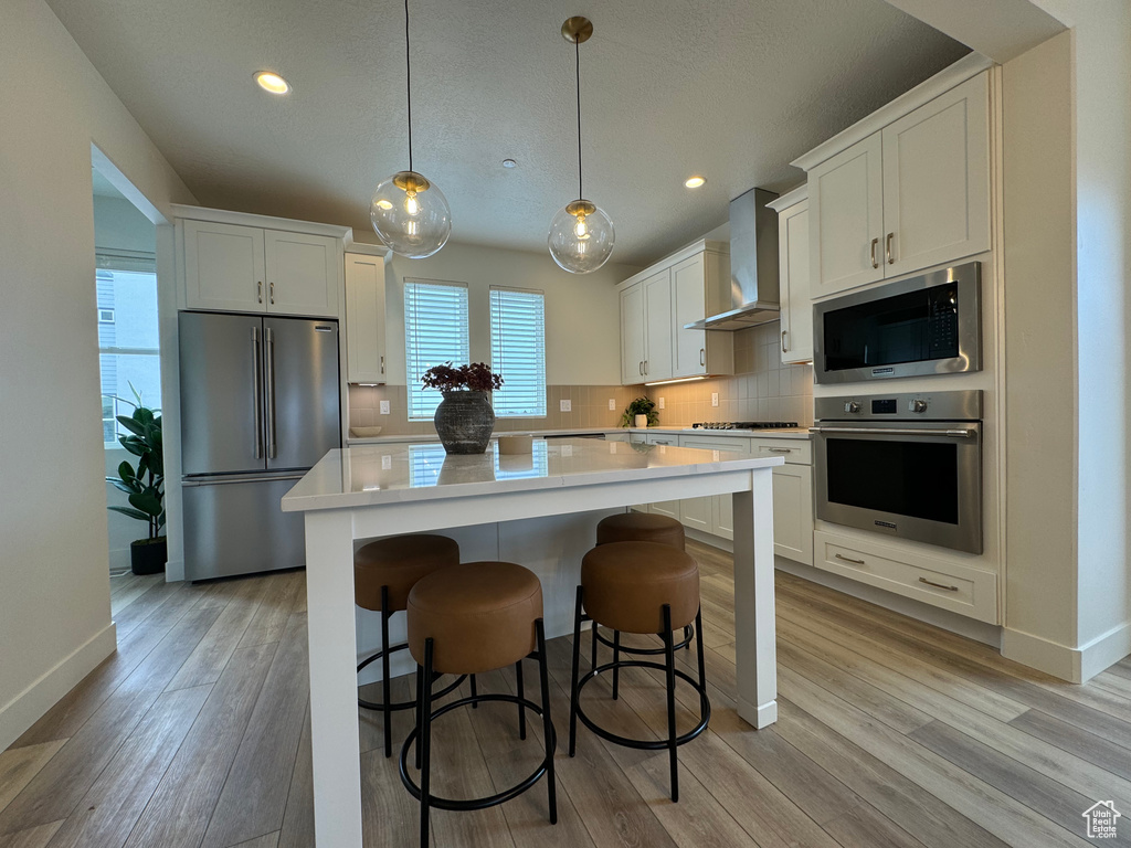 Kitchen featuring a kitchen island, wall chimney range hood, light hardwood / wood-style floors, backsplash, and appliances with stainless steel finishes