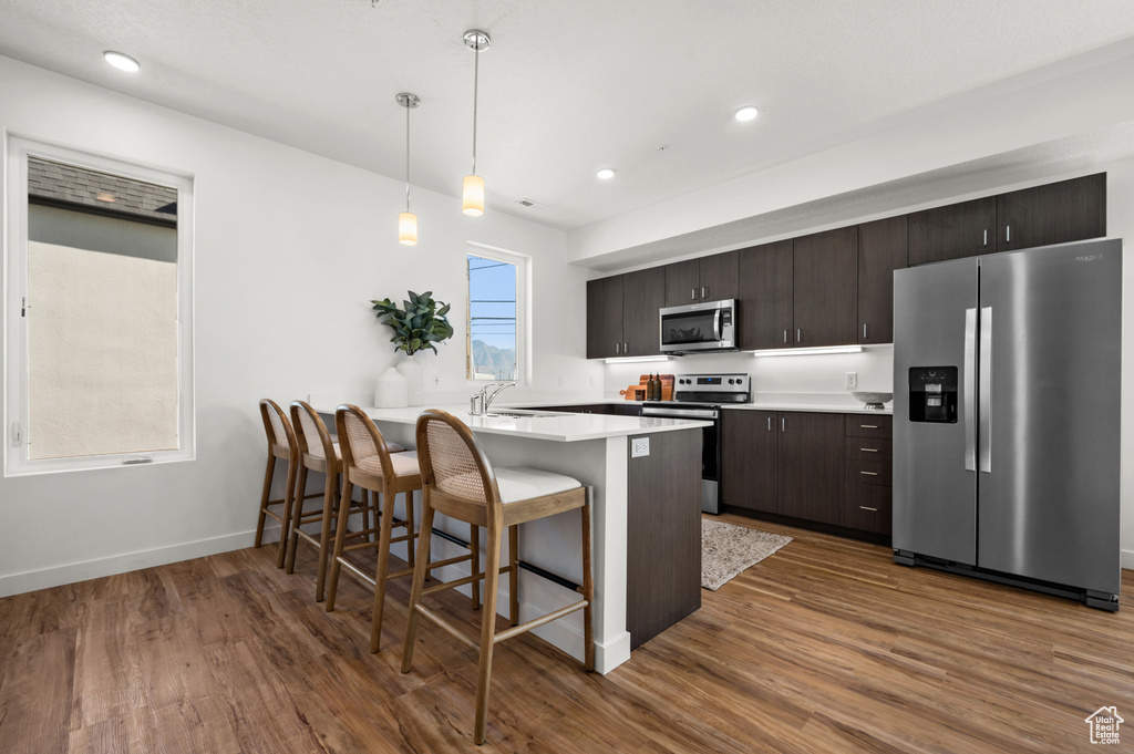 Kitchen with a kitchen breakfast bar, stainless steel appliances, dark hardwood / wood-style floors, dark brown cabinets, and pendant lighting