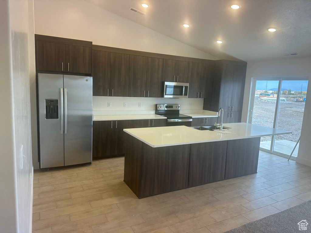 Kitchen featuring appliances with stainless steel finishes, sink, lofted ceiling, dark brown cabinetry, and an island with sink