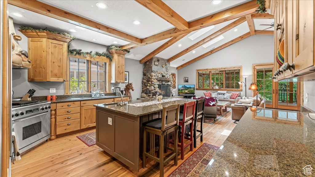 Kitchen featuring a stone fireplace, light hardwood / wood-style floors, an island with sink, beamed ceiling, and high end stove