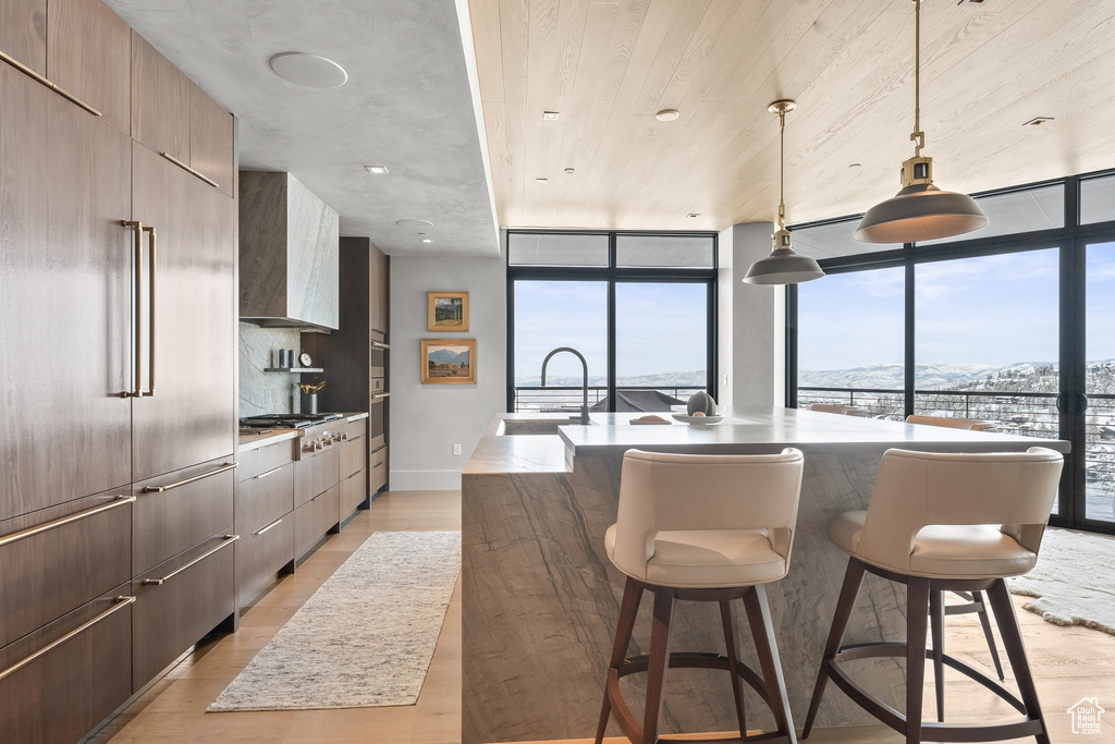 Kitchen with floor to ceiling windows, a kitchen island with sink, light hardwood / wood-style flooring, a kitchen breakfast bar, and wall chimney range hood