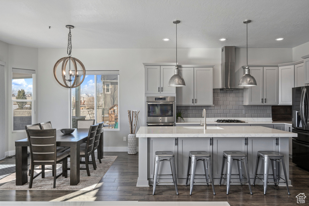 Kitchen featuring a breakfast bar, wall chimney range hood, decorative light fixtures, a center island with sink, and gray cabinetry