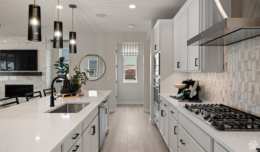 Kitchen with appliances with stainless steel finishes, wall chimney range hood, white cabinets, pendant lighting, and light wood-type flooring