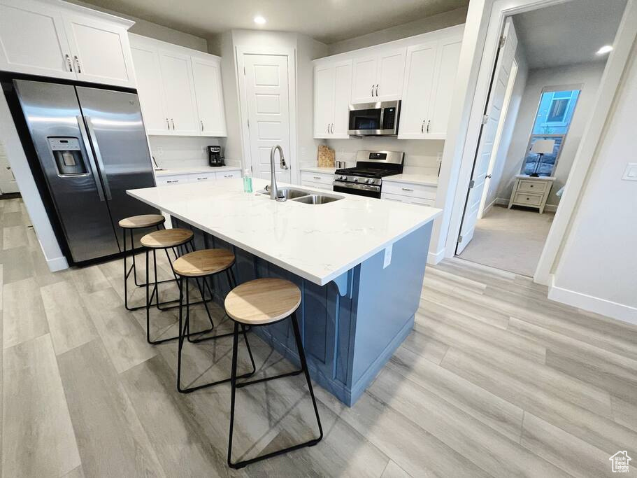 Kitchen with stainless steel appliances, white cabinetry, light wood-type flooring, an island with sink, and a kitchen bar