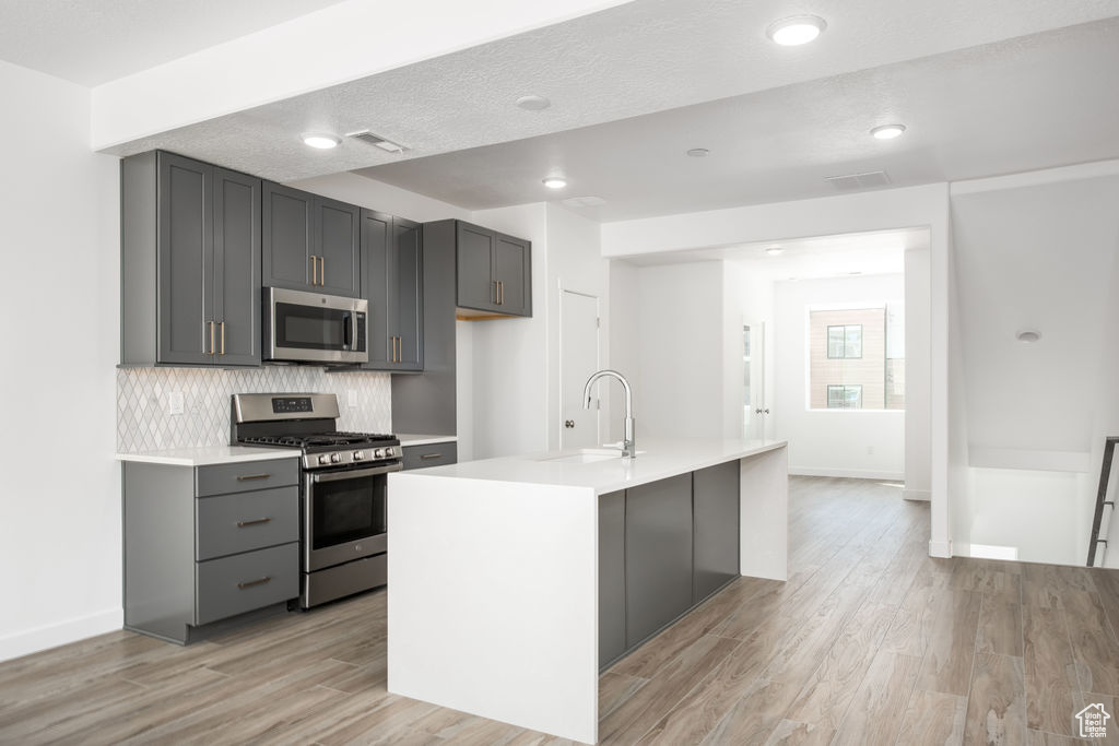 Kitchen featuring stainless steel appliances, light wood-type flooring, a kitchen island with sink, and gray cabinetry
