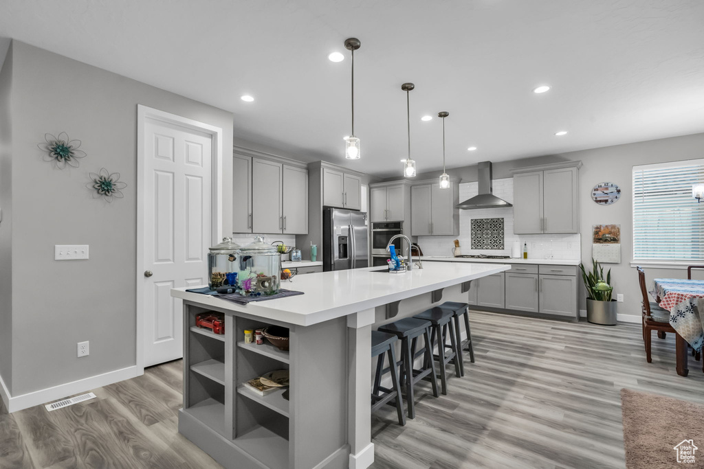 Kitchen with an island with sink, appliances with stainless steel finishes, gray cabinets, light hardwood / wood-style floors, and wall chimney range hood