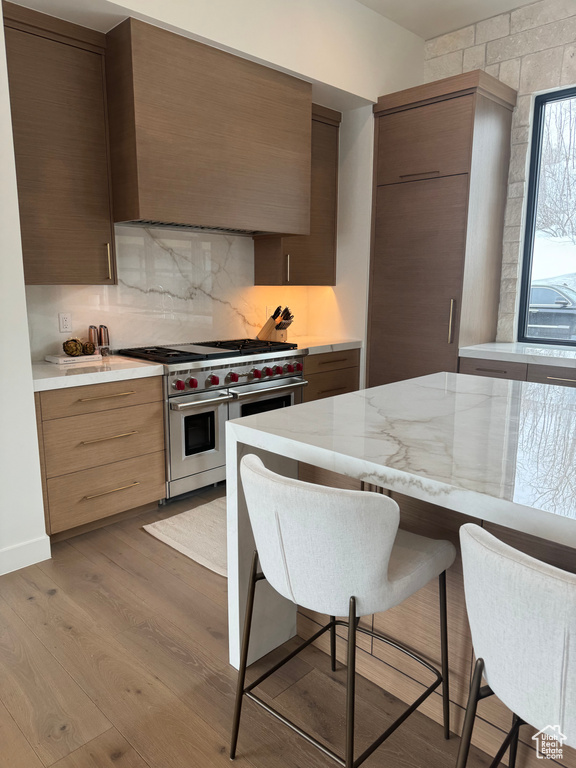 Kitchen featuring wall chimney exhaust hood, double oven range, a kitchen bar, light hardwood / wood-style floors, and tasteful backsplash