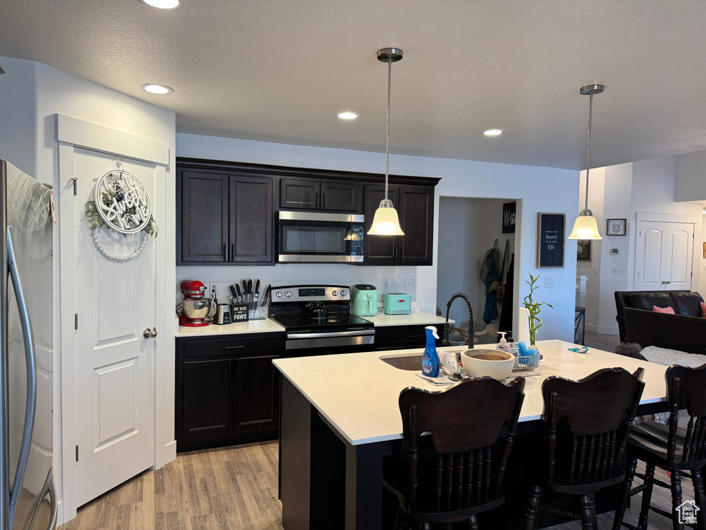 Kitchen featuring a center island with sink, stainless steel appliances, hanging light fixtures, and light hardwood / wood-style flooring