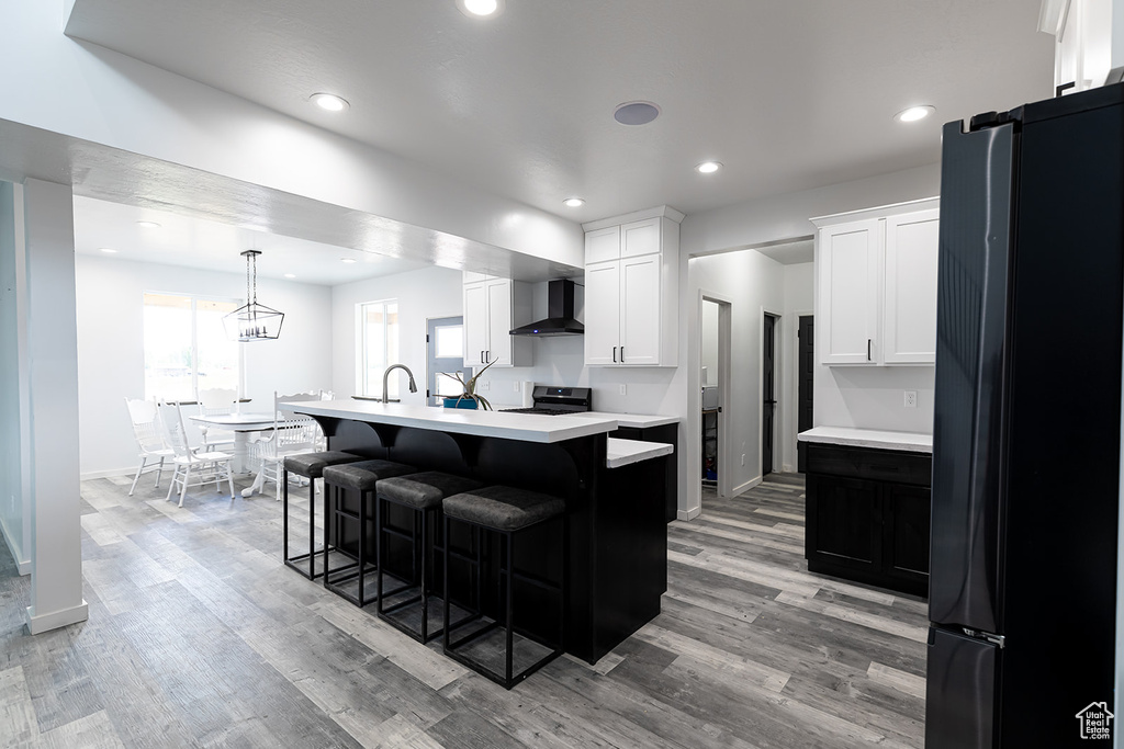 Kitchen featuring a kitchen island with sink, wall chimney range hood, light hardwood / wood-style floors, decorative light fixtures, and black fridge
