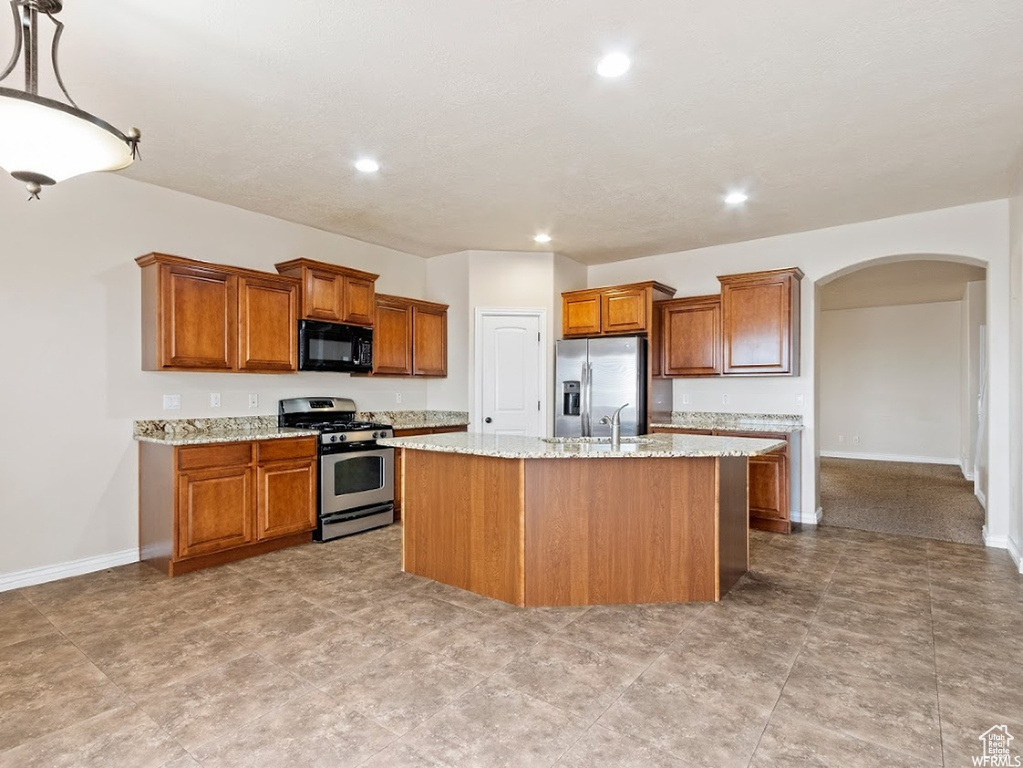Kitchen featuring appliances with stainless steel finishes, a center island with sink, light tile floors, pendant lighting, and light stone countertops