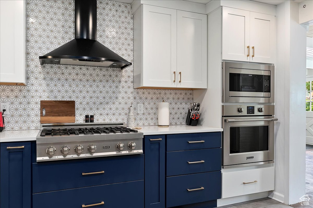 Kitchen featuring white cabinetry, blue cabinets, wall chimney exhaust hood, backsplash, and stainless steel appliances