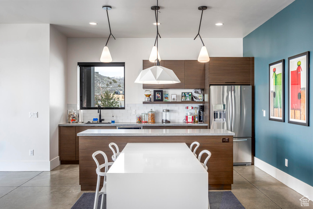 Kitchen with backsplash, stainless steel fridge, concrete flooring, hanging light fixtures, and a kitchen island