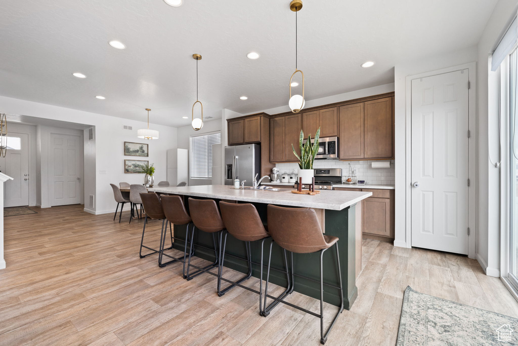 Kitchen featuring an island with sink, light hardwood / wood-style flooring, backsplash, stainless steel appliances, and pendant lighting