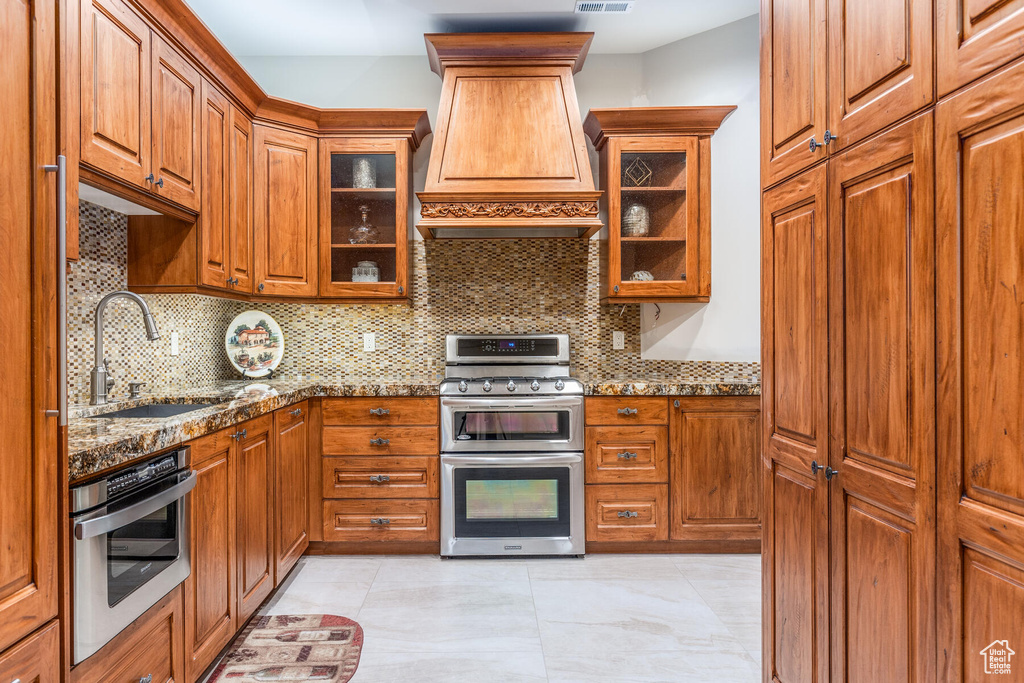 Kitchen with custom range hood, stainless steel appliances, light stone counters, sink, and tasteful backsplash