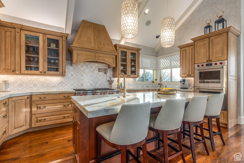 Kitchen featuring dark wood-type flooring, premium range hood, a kitchen island with sink, pendant lighting, and tasteful backsplash