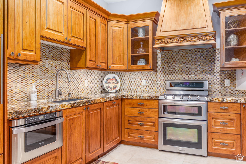 Kitchen featuring light tile flooring, tasteful backsplash, custom exhaust hood, appliances with stainless steel finishes, and sink