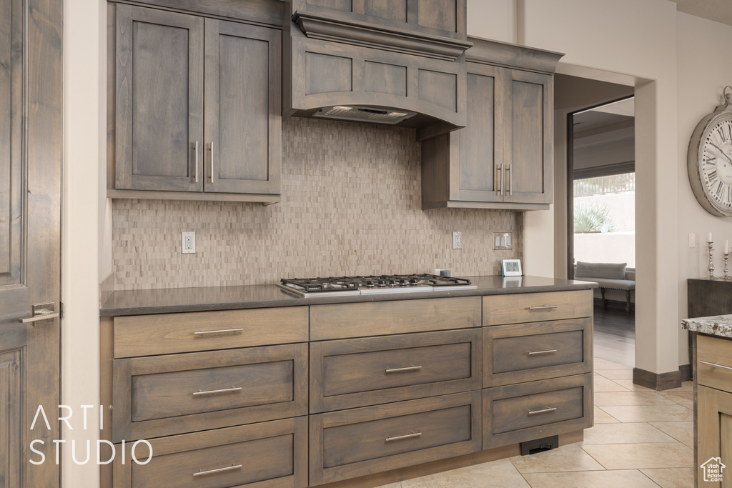 Kitchen featuring stainless steel gas stovetop, custom range hood, tasteful backsplash, light tile flooring, and dark brown cabinets